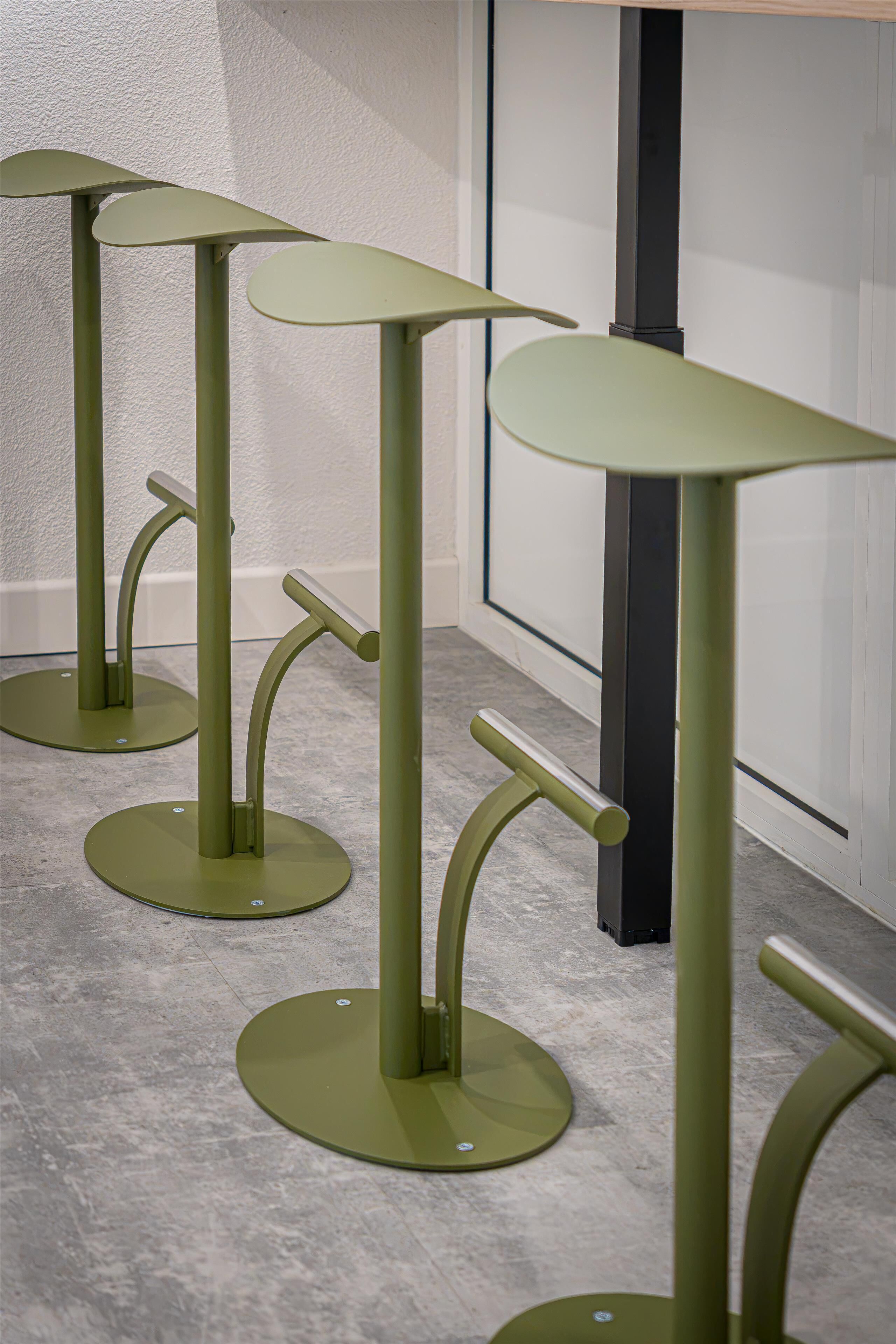 A series of green, modern, minimalist tables with curved legs and round tops, arranged in a row against a white wall with a dark column in the background.
