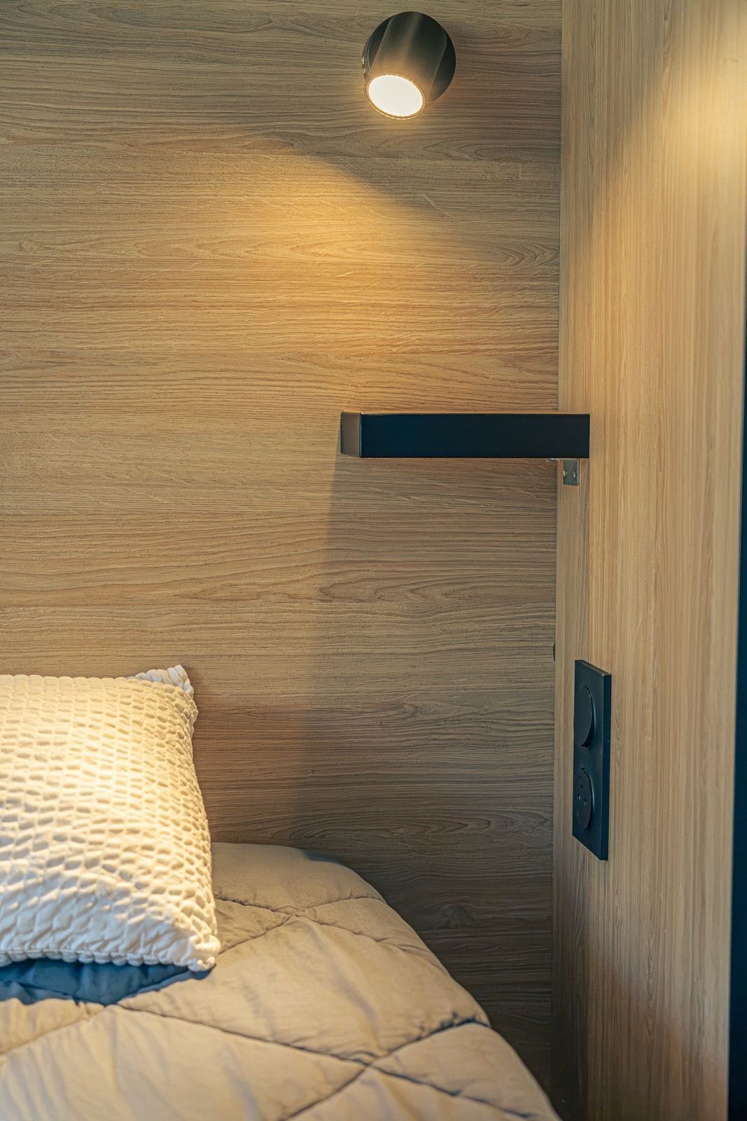 Una cama con un almohadón texturizado, un muro de madera y una lámpara en el techo.