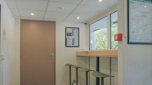 Un pasillo con una puerta cerrada, un tablón de anuncios con avisos, una ventana con vistas y un detector de incendios rojo.