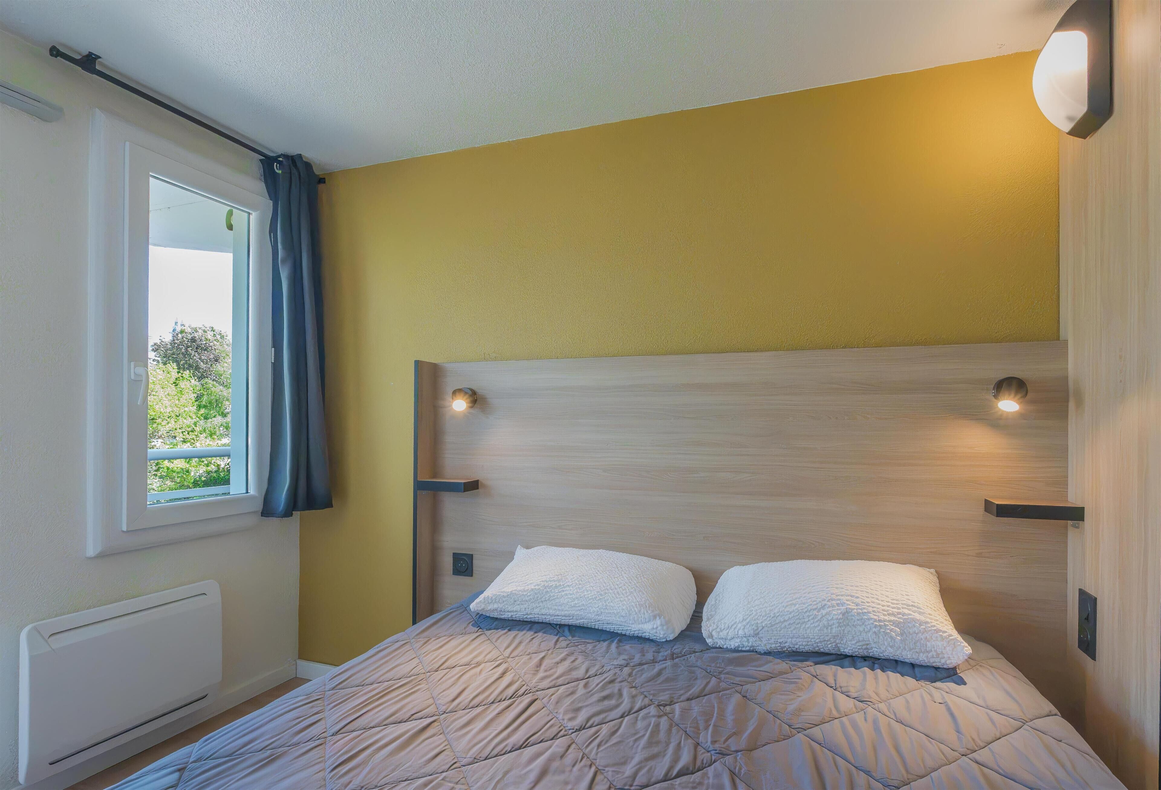 Une chambre à coucher avec un lit, une fenêtre avec des rideaux et un luminaire fixé au mur.