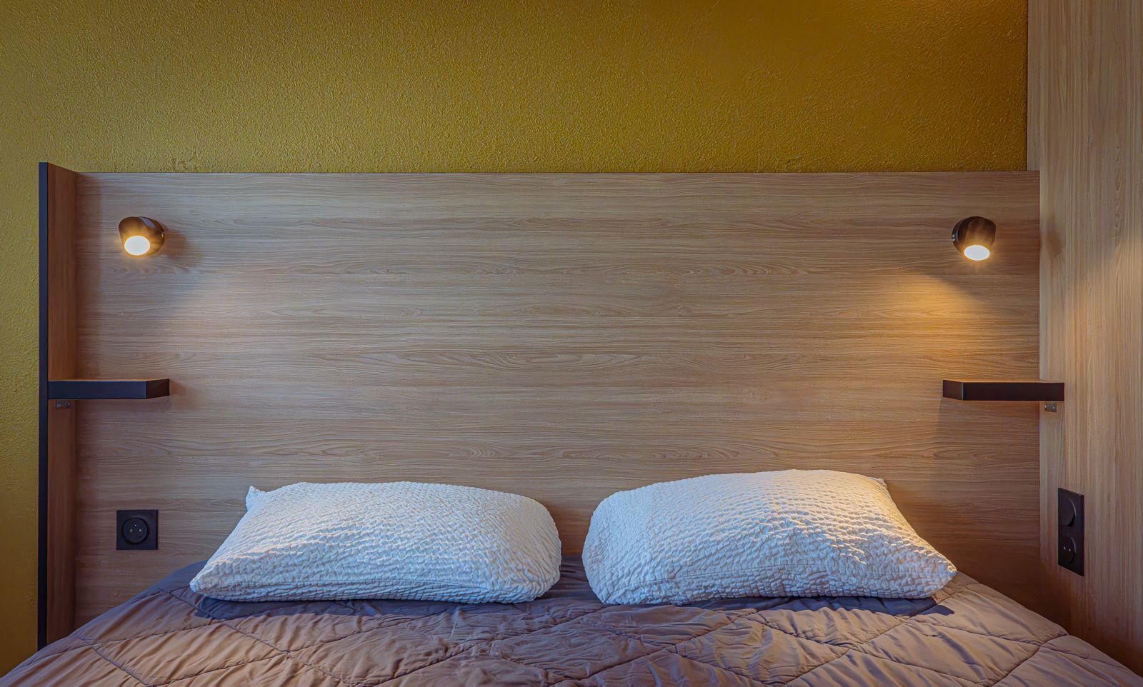 Un lit avec une tête de lit en bois, deux oreillers et deux lampes fixées au mur.