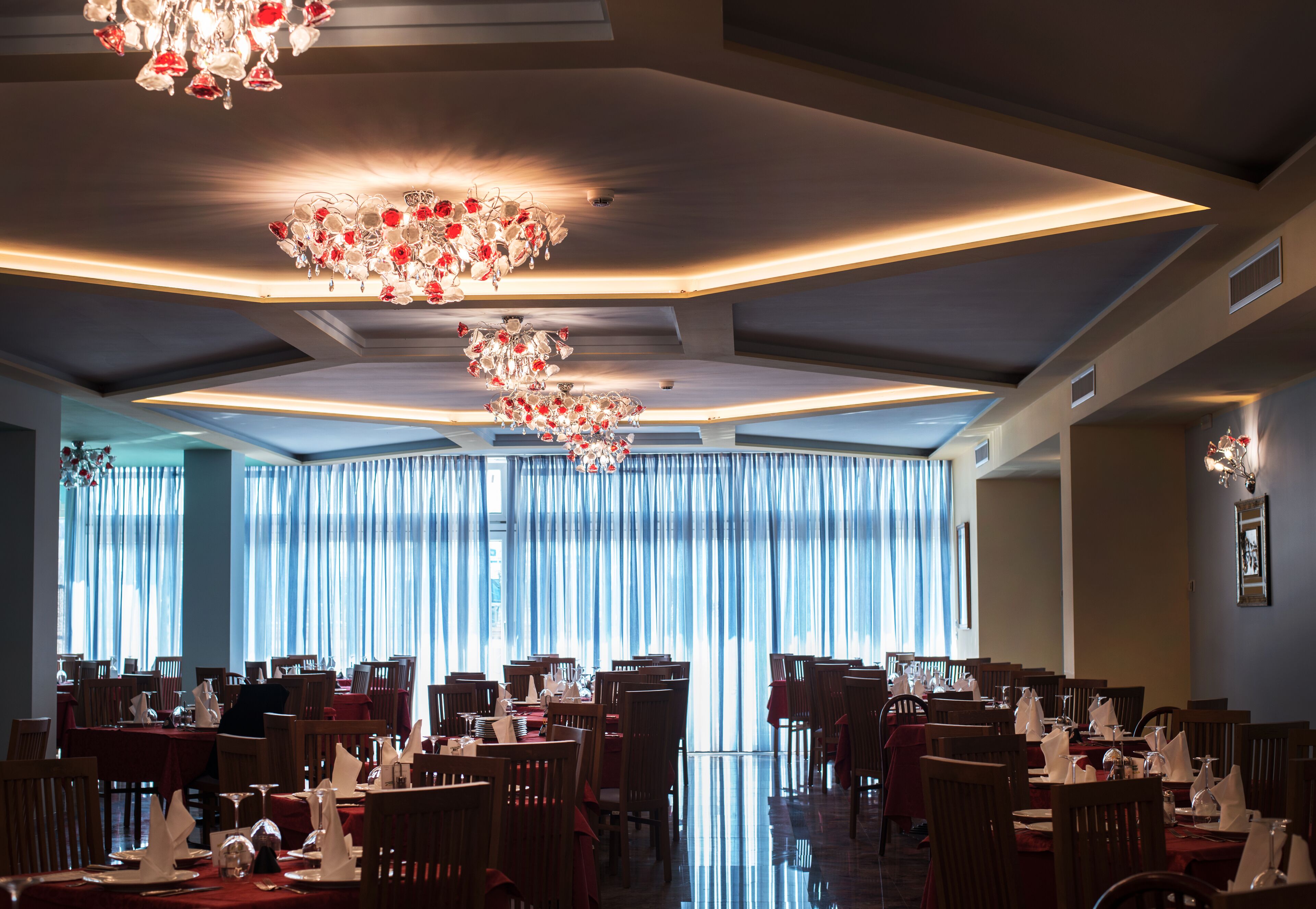 Un restaurante con varias mesas y sillas, un candelabro y ventanas amplias con cortinas.