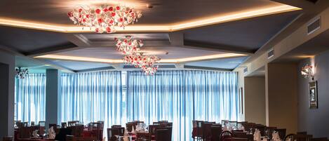 A restaurant with multiple tables and chairs, a chandelier, and large windows with curtains.