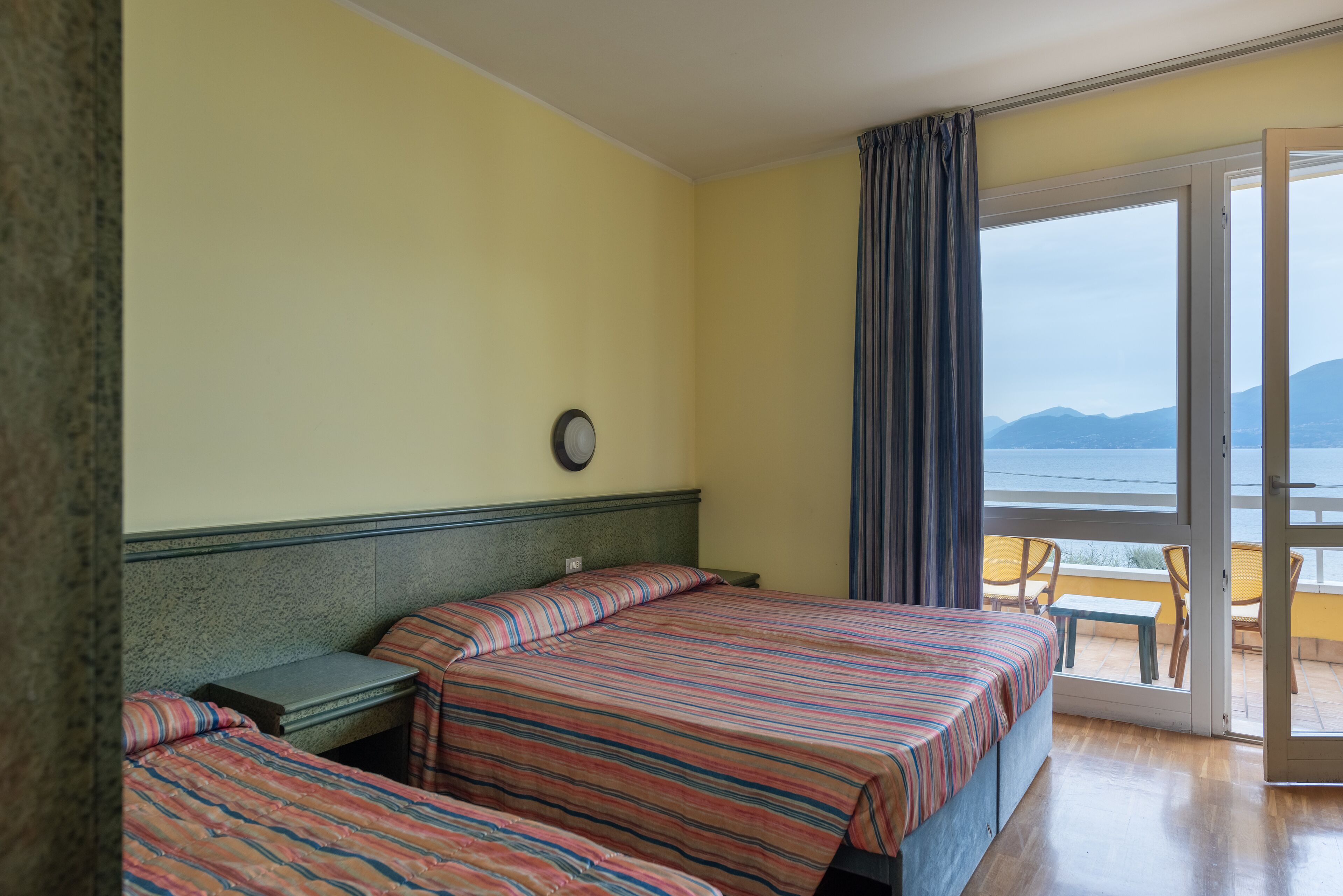A hotel room with two beds, a window with a view of the sea, and a balcony with chairs.