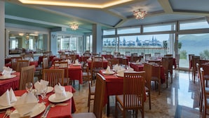 A restaurant with tables set for dining, large windows offering a view of a cityscape and mountains, and a modern chandelier. - Hotel Internazionale (Torri del Benaco)