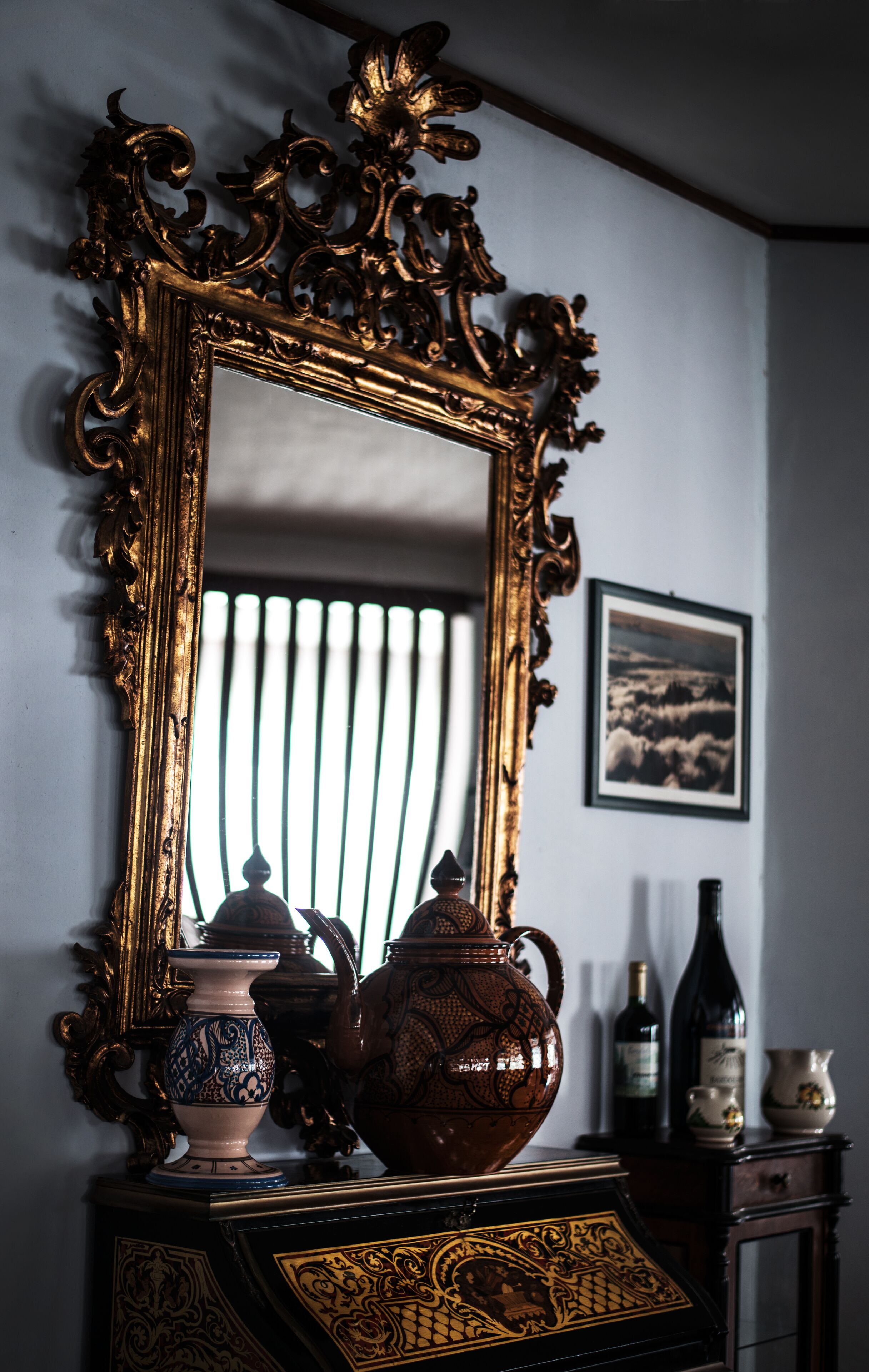 A room with a large ornate mirror, a vase, a teapot, and a wine bottle on a cabinet.