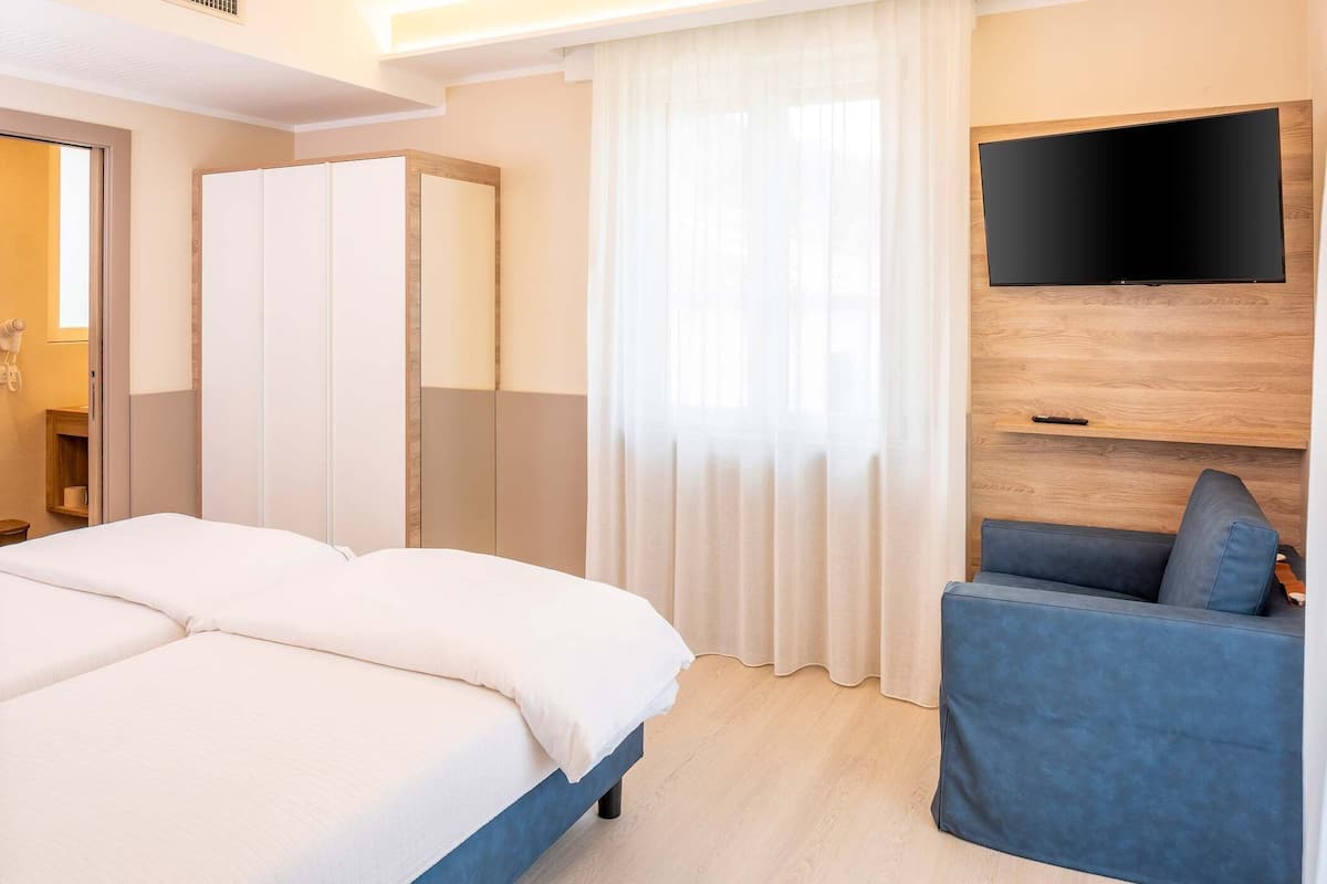 A hotel room with a bed, a TV mounted on the wall, a seating area, and a window with curtains.