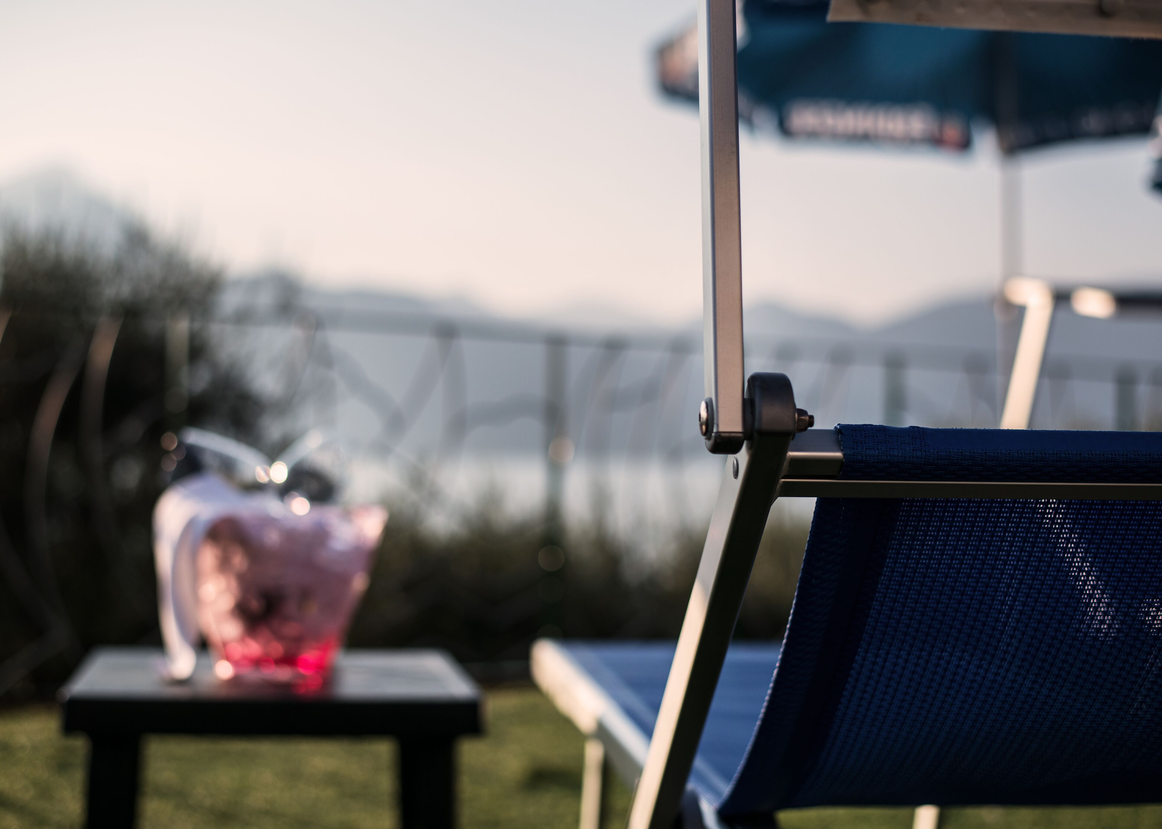 A blue outdoor chair with a metal frame, a small table with a pink object, and a blue umbrella in the background.