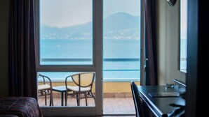 A hotel room with a bed, a desk, and a chair. There is a large window with a view of the sea and mountains. - Hotel Internazionale (Torri del Benaco)