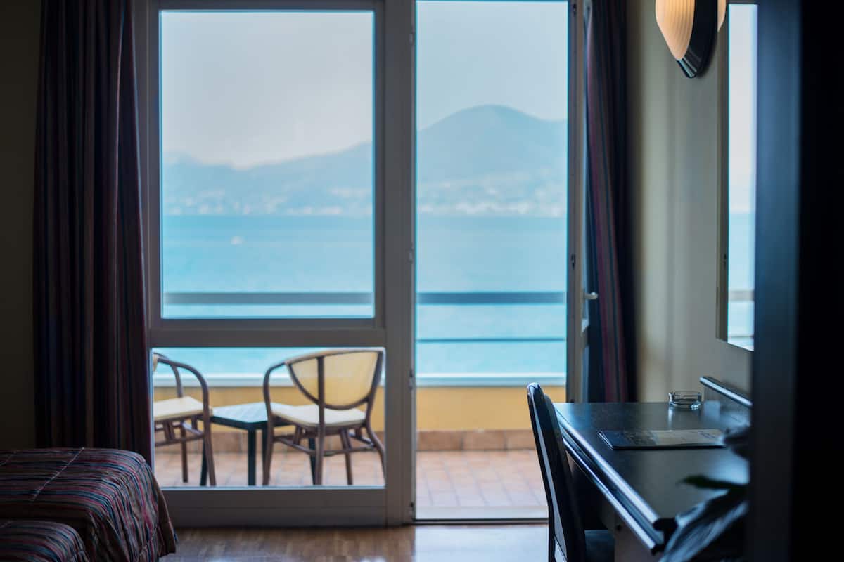 A hotel room with a bed, a desk, and a chair. There is a large window with a view of the sea and mountains.