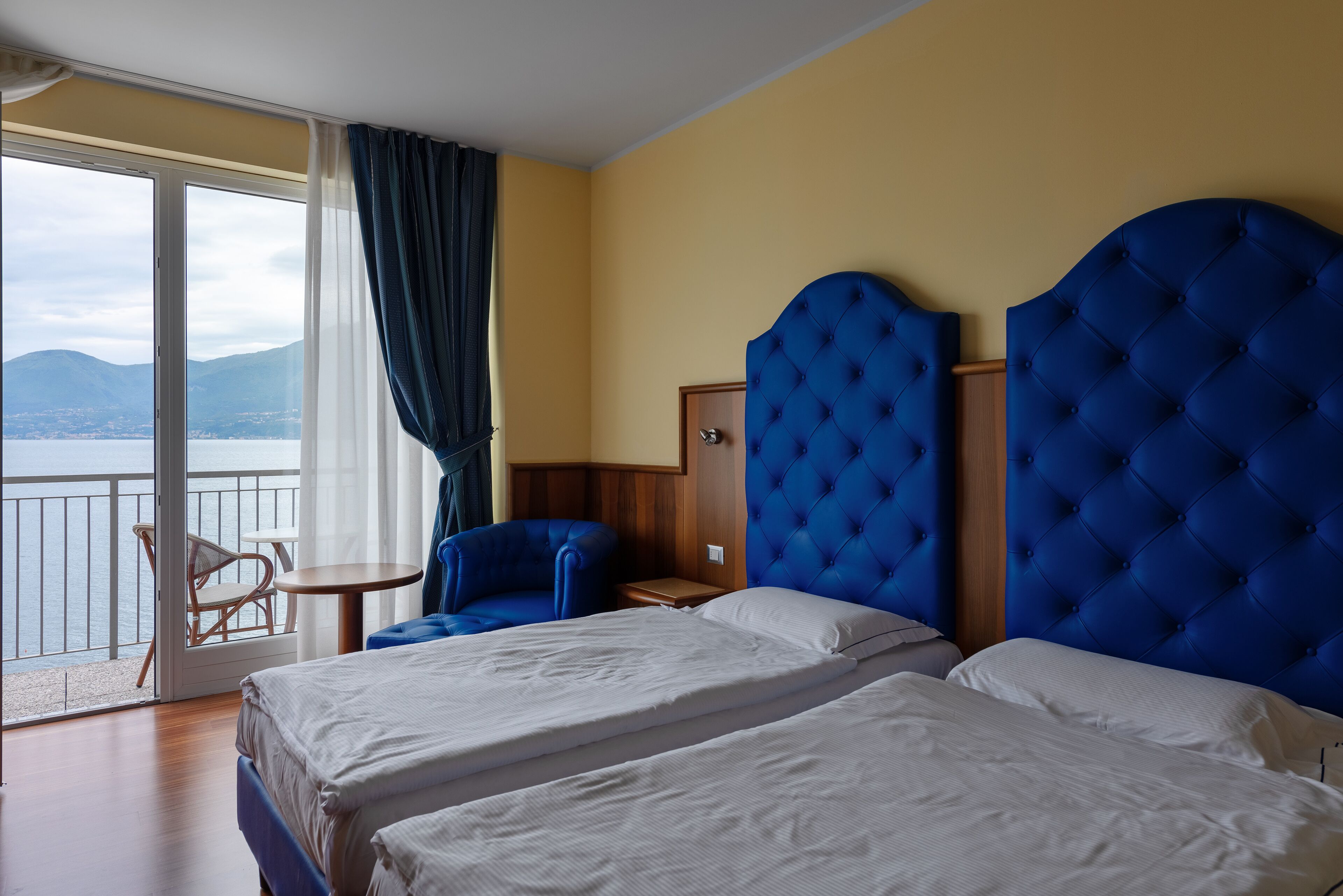 Une chambre d’hôtel avec deux lits, un balcon donnant sur la mer et un fauteuil bleu recouvert de tissu.