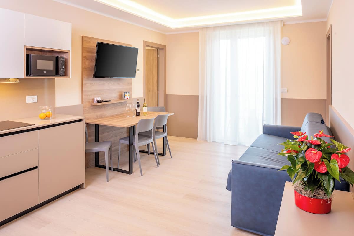 A modern living room with a dining area, a flat-screen TV mounted on the wall, a kitchen with a microwave, and a potted plant.