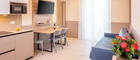 Una sala de estar moderna con comedor, televisor de pantalla plana en la pared, cocina con microondas y una planta en maceta.