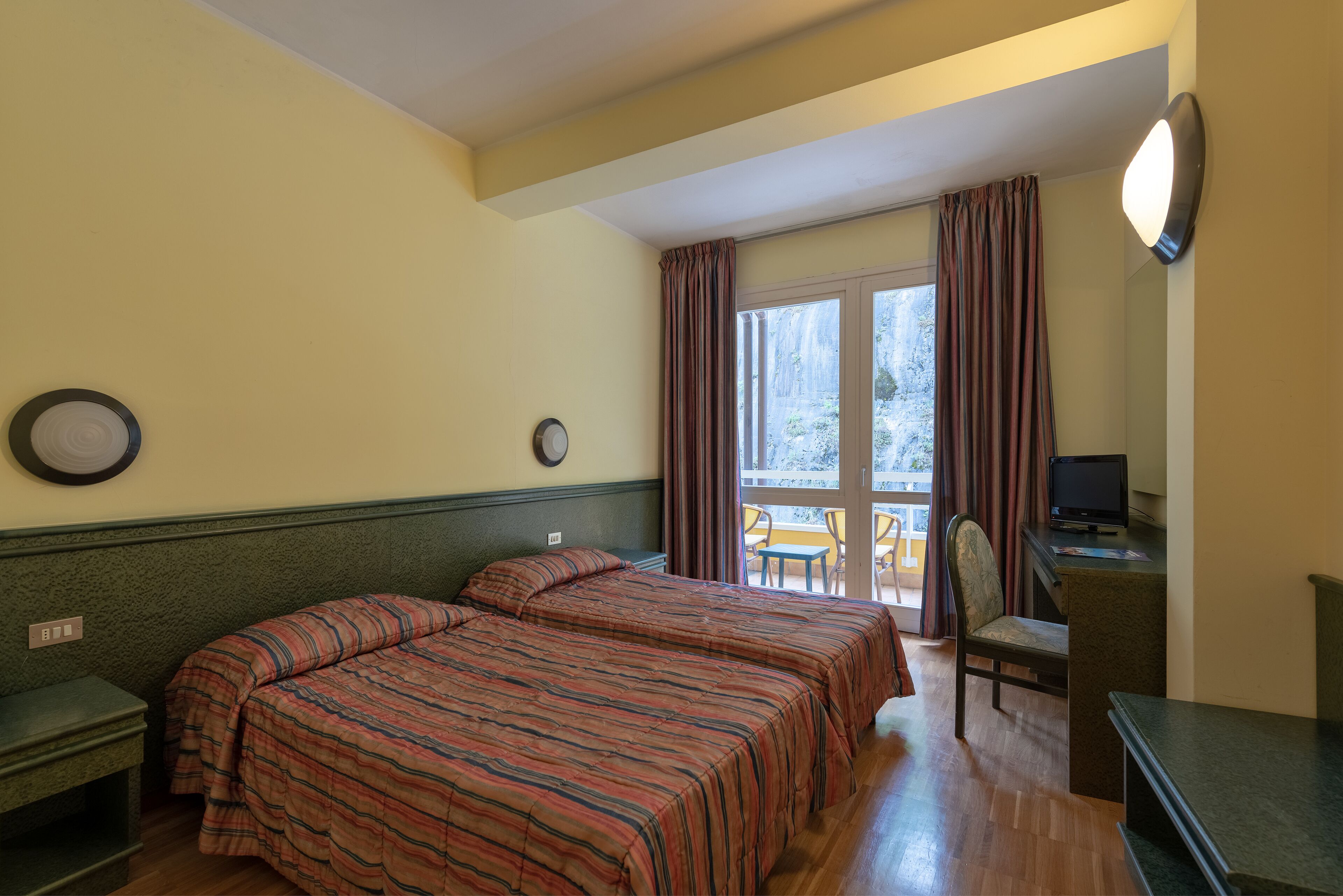 A hotel room with two beds, a desk, a chair, a TV, and a balcony with a view.