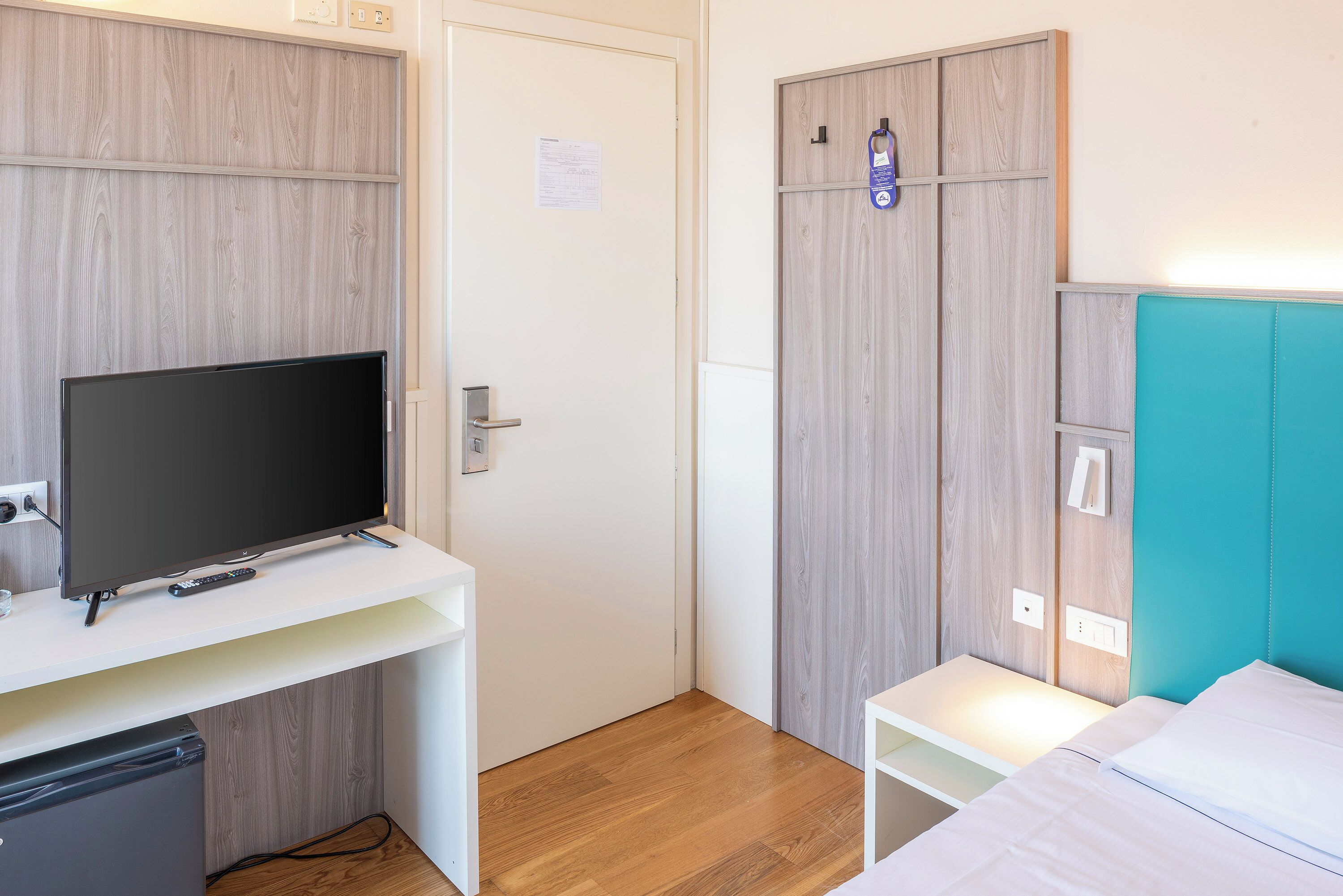 Una habitación compacta con un televisor sobre un soporte blanco, un armario de madera y una cama con cabecera color turquesa.