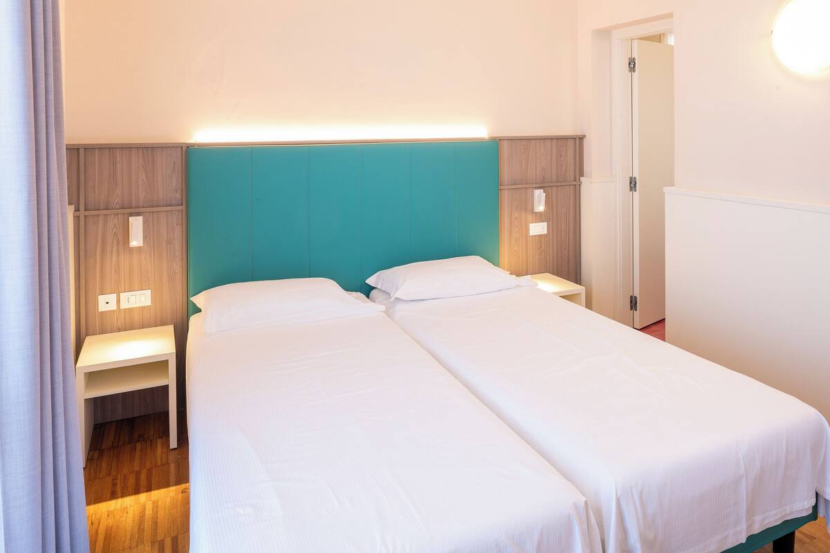 A hotel room with two single beds, a nightstand, a wall-mounted light, and a wooden headboard.
