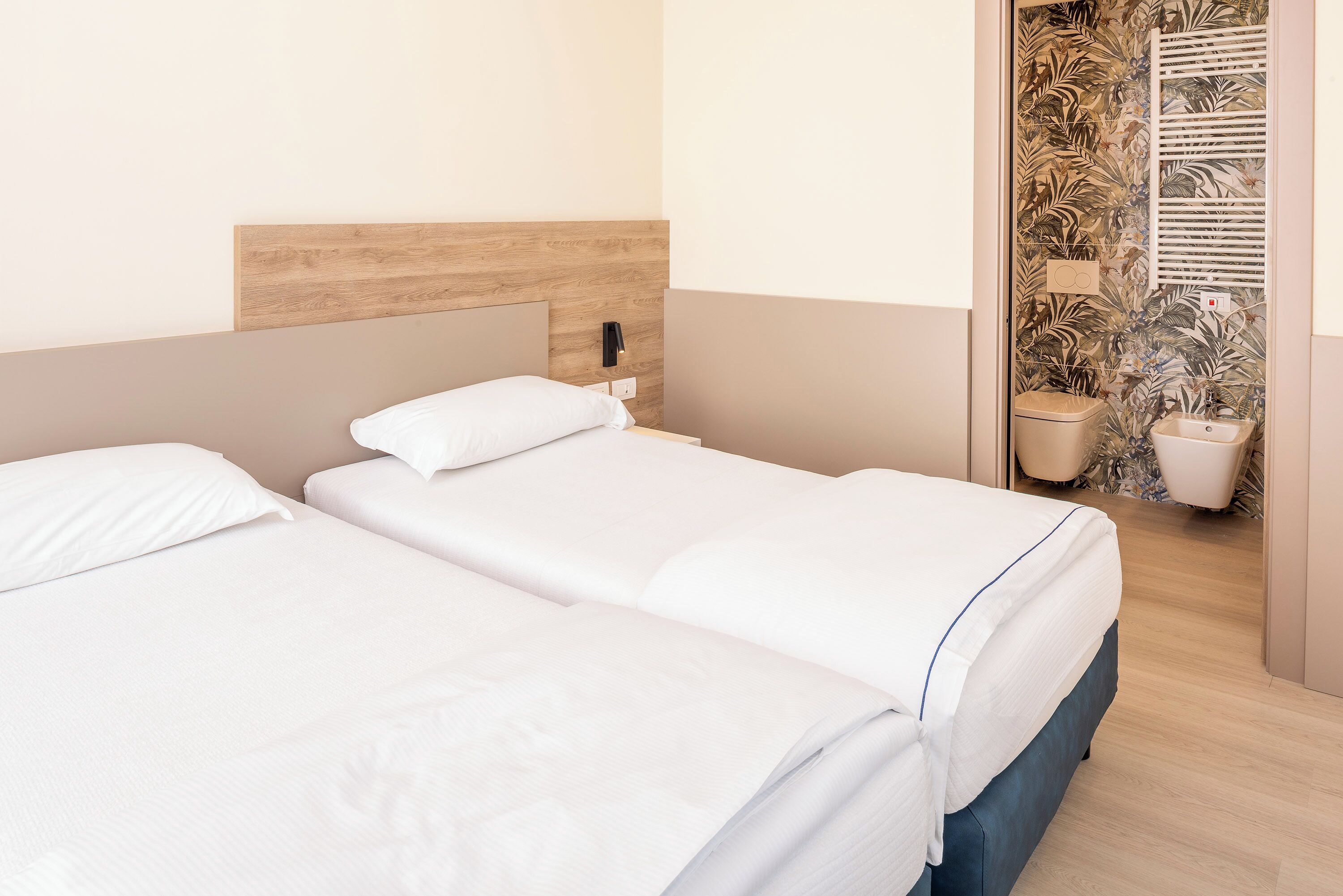 A hotel room with two beds, a wooden headboard, a small bathroom with a shower, and a wall with a leaf pattern.