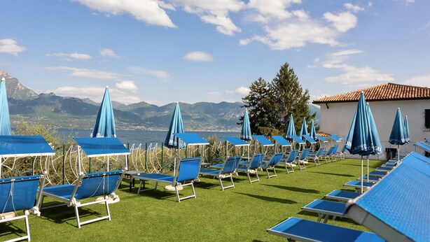 A row of blue lounge chairs with umbrellas on a grassy area, overlooking a body of water and mountains. - Hotel Internazionale (Torri del Benaco)