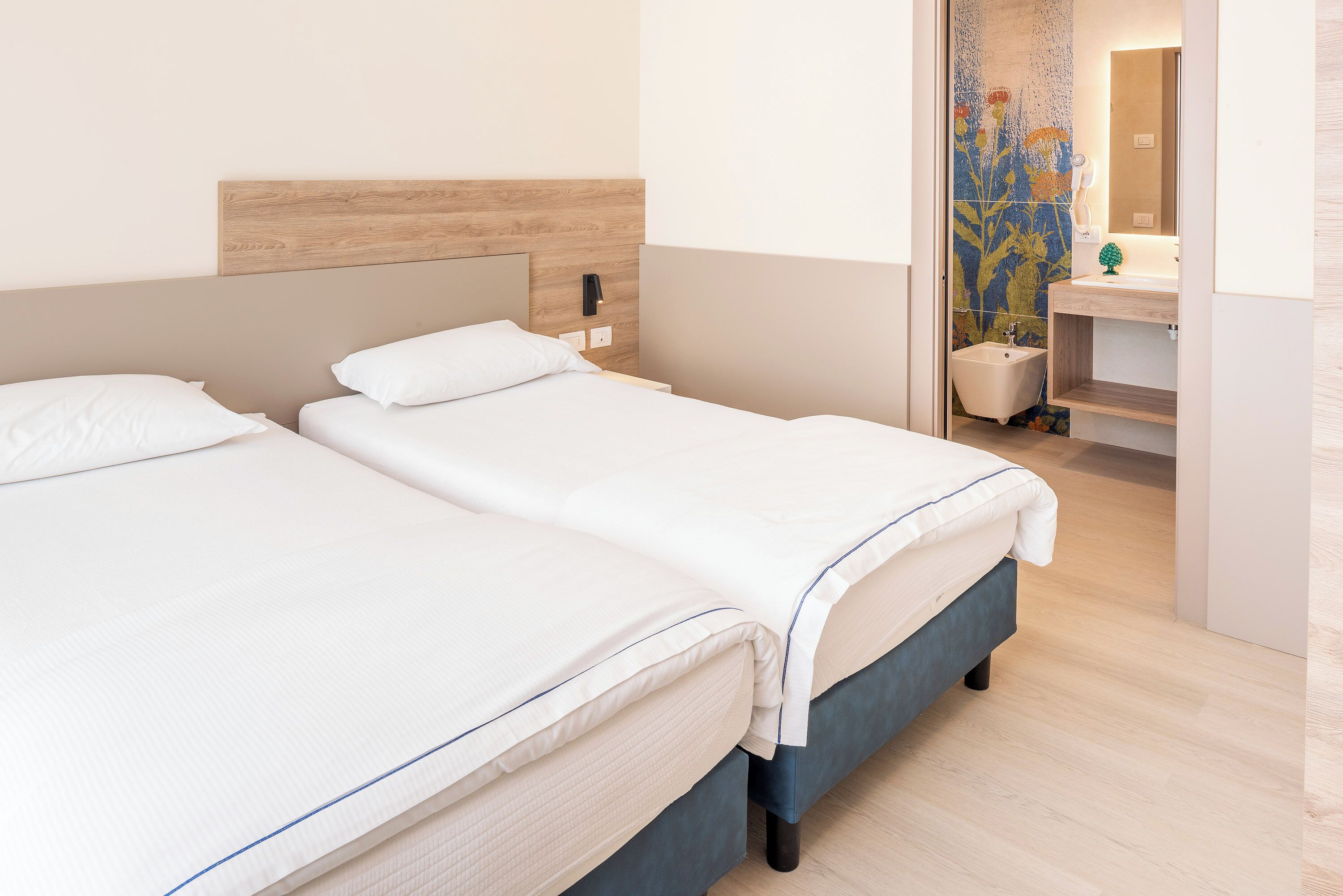 A hotel room with two beds, a wooden headboard, a bathroom with a floral tile backsplash, and a small sink.