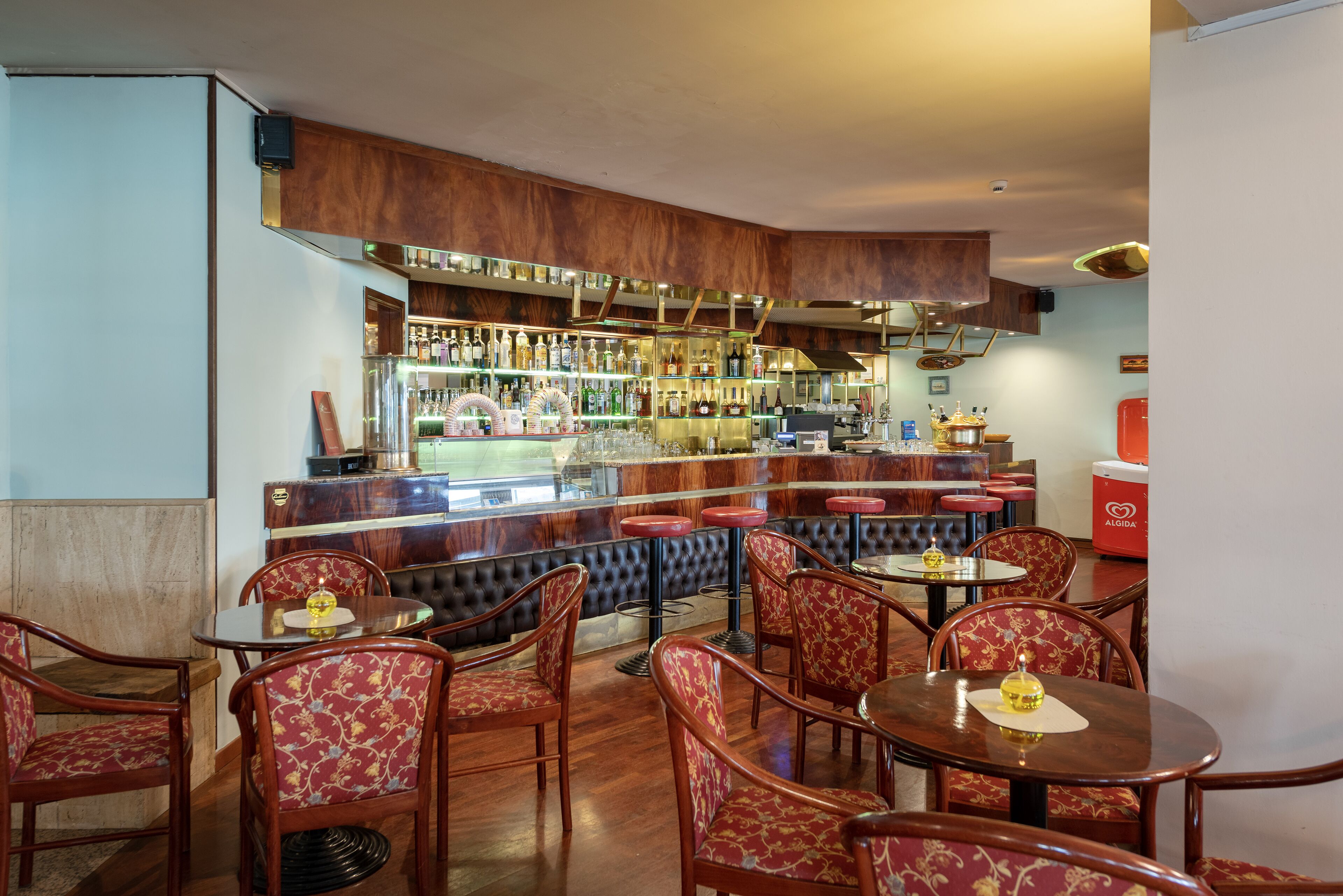 A bar area with wooden bar counters, multiple stools, and a variety of bottles and glasses on shelves.