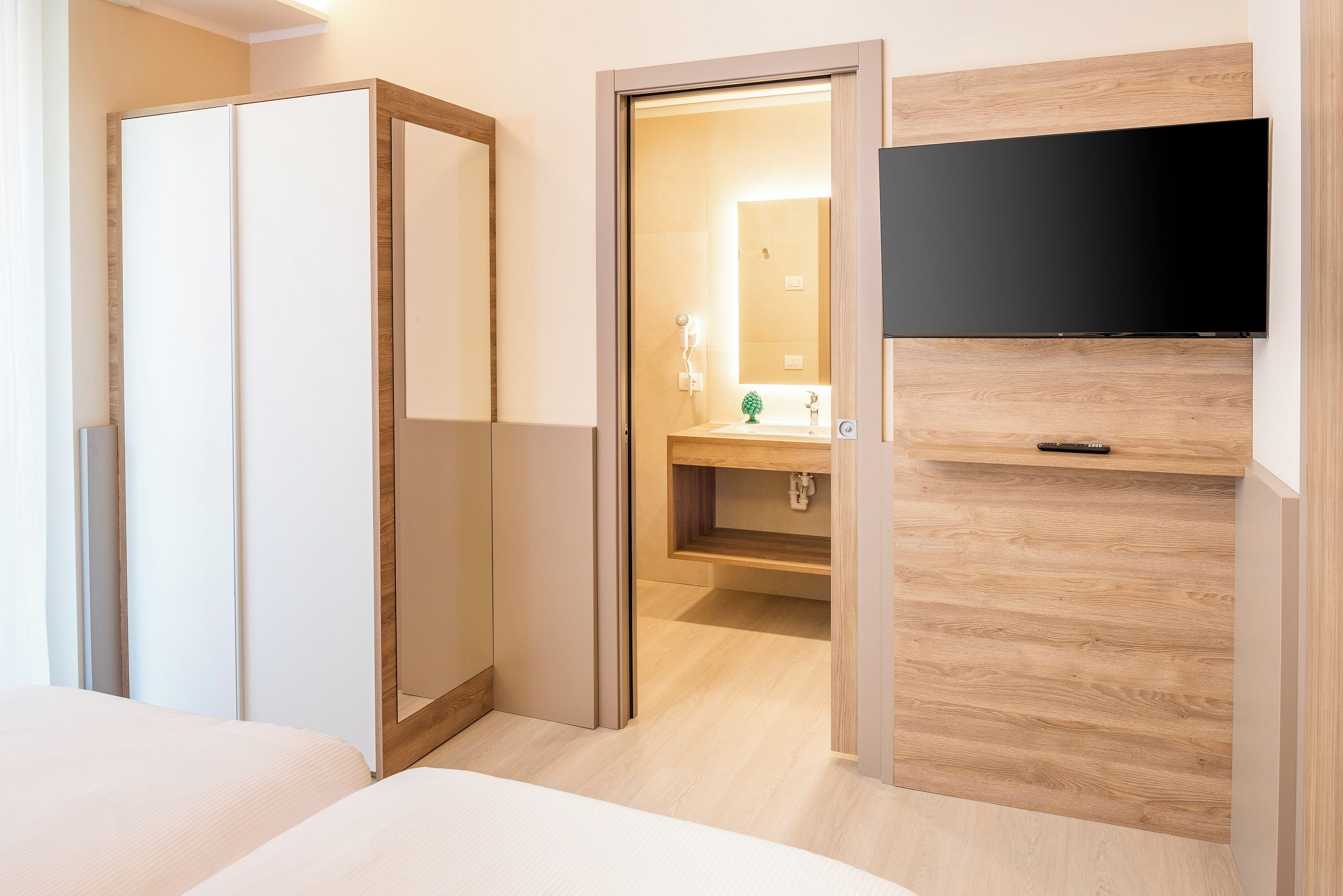A hotel room with a bed, a TV mounted on the wall, a wooden wardrobe, and a bathroom visible through an open door.
