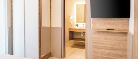 A hotel room with a bed, a TV mounted on the wall, a wooden wardrobe, and a bathroom visible through an open door.