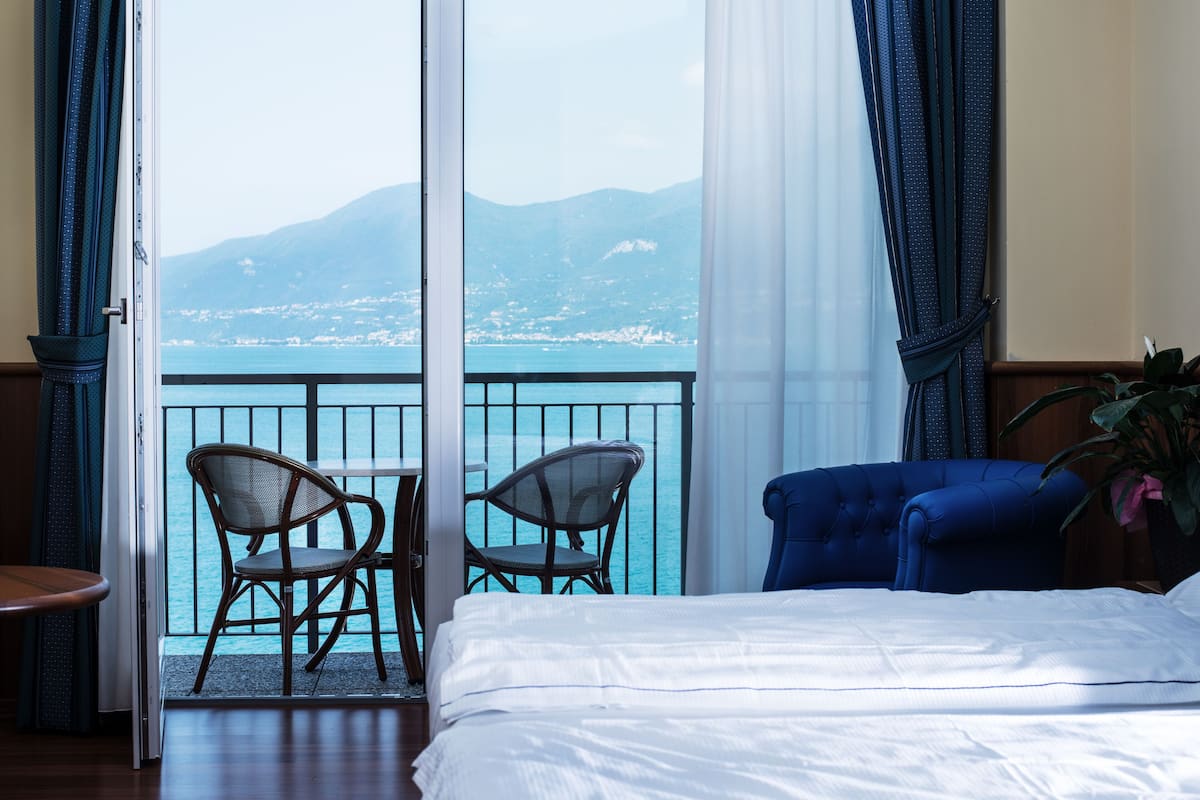 A hotel room with a bed, two wicker chairs, a small table, and a view of the sea and mountains.