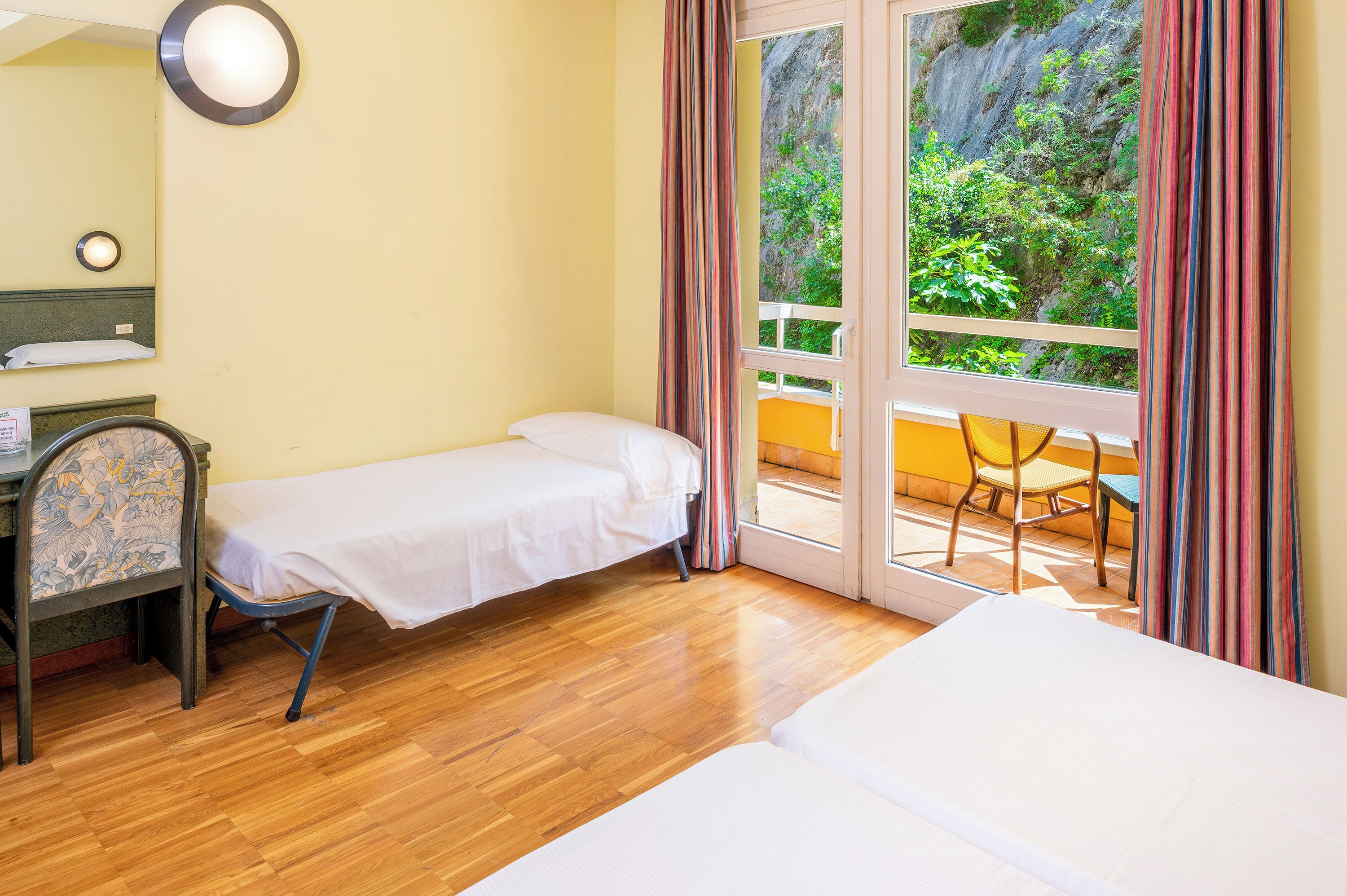 A room with a bed, a desk, and a chair. There is a window with curtains, and a view of greenery outside.