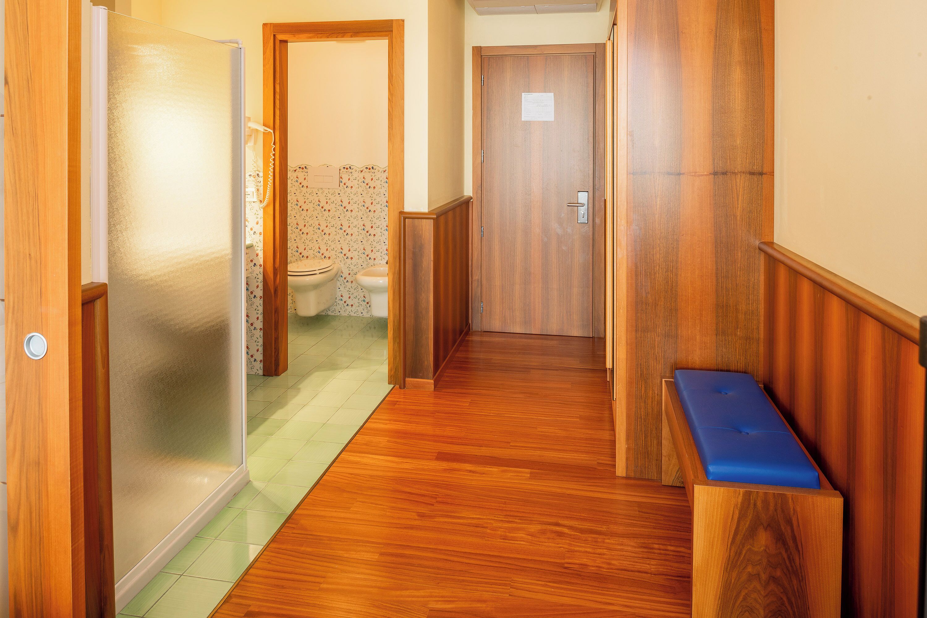 A hallway with wooden flooring, a bench, and a door leading to a bathroom with a toilet and patterned wallpaper.