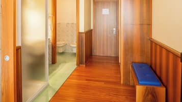 Un pasillo con suelo de madera, un banco y una puerta que da a un baño con inodoro y papel tapiz estampado.