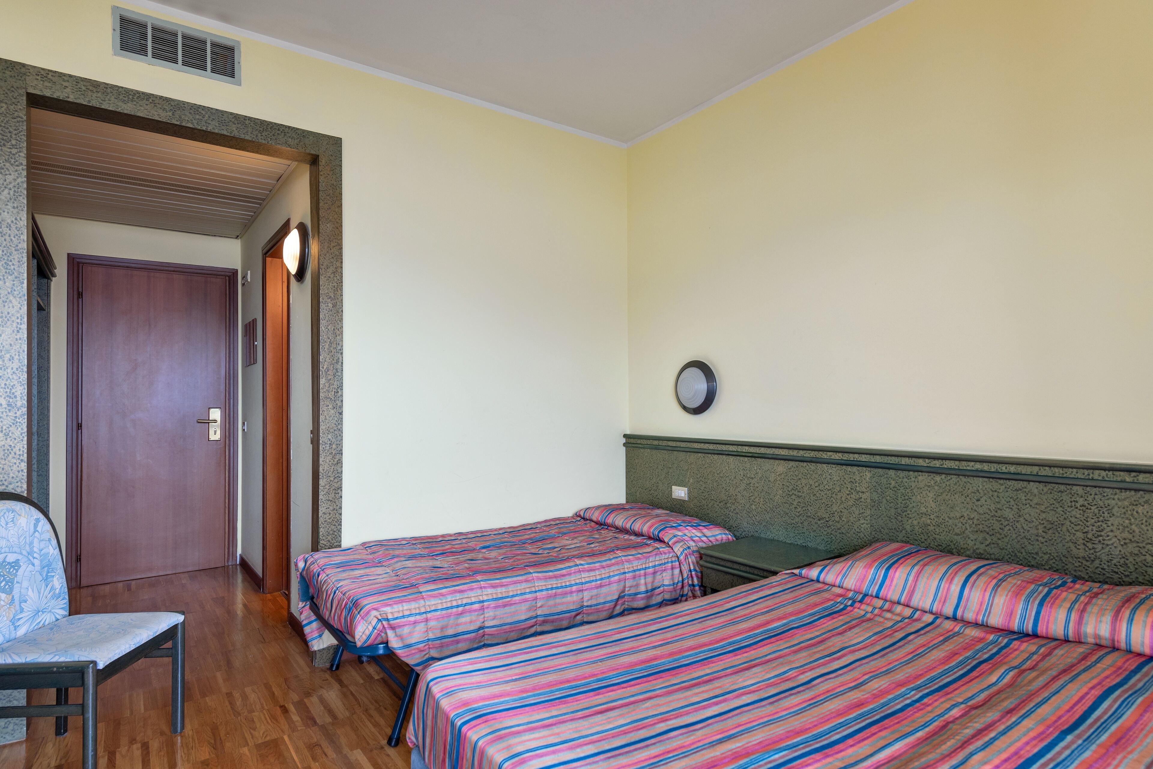 Een kamer met twee bedden, een stoel, een deur en een wandklok.