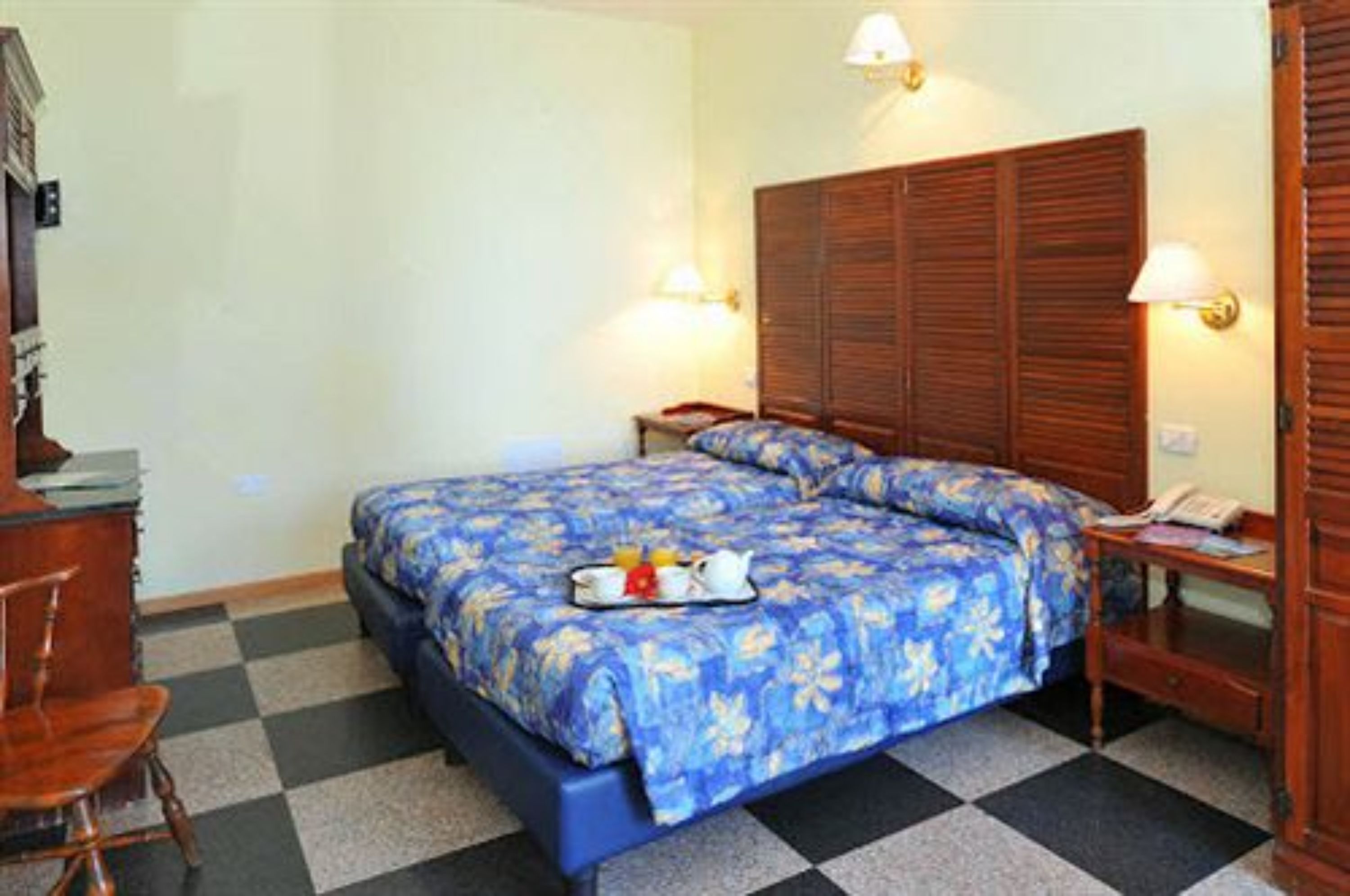 A hotel room with a checkered floor, a bed with a floral bedspread, two bedside lamps, a wooden wardrobe, and a small table with a tray of breakfast items.