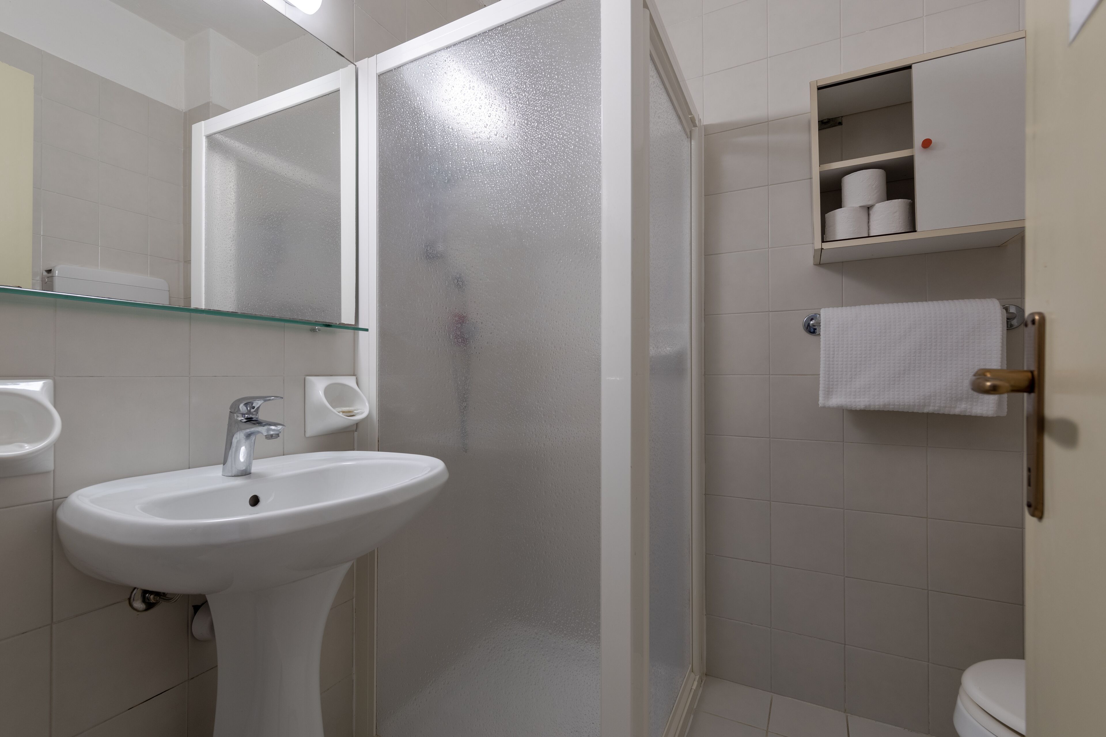 A bathroom with a white sink, a glass shower enclosure, a toilet, and a small cabinet with toiletries.
