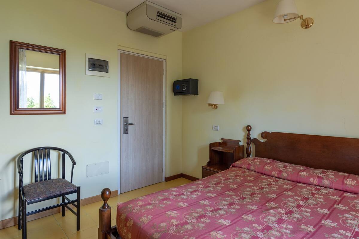 Un dormitorio con cama, una silla, una mesita de noche, lámpara, armario y unidad de aire acondicionado.