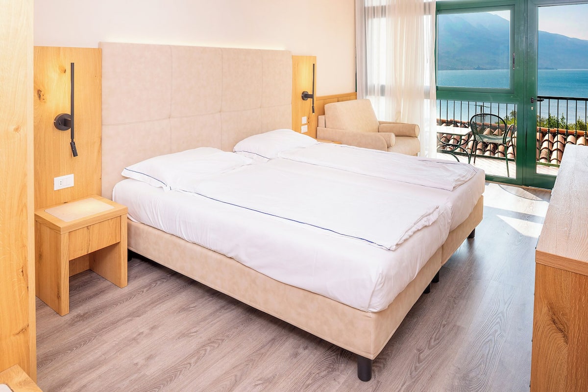 Una cama doble con cabecera de madera, una mesita de noche y vistas a un balcón con una silla.