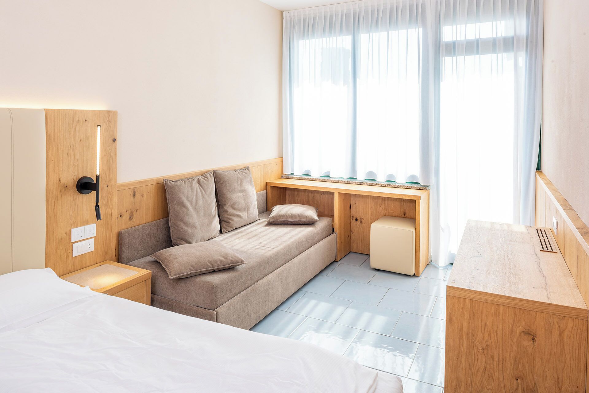 A compact hotel room with a bed, a sofa, a desk, and a window with sheer curtains.