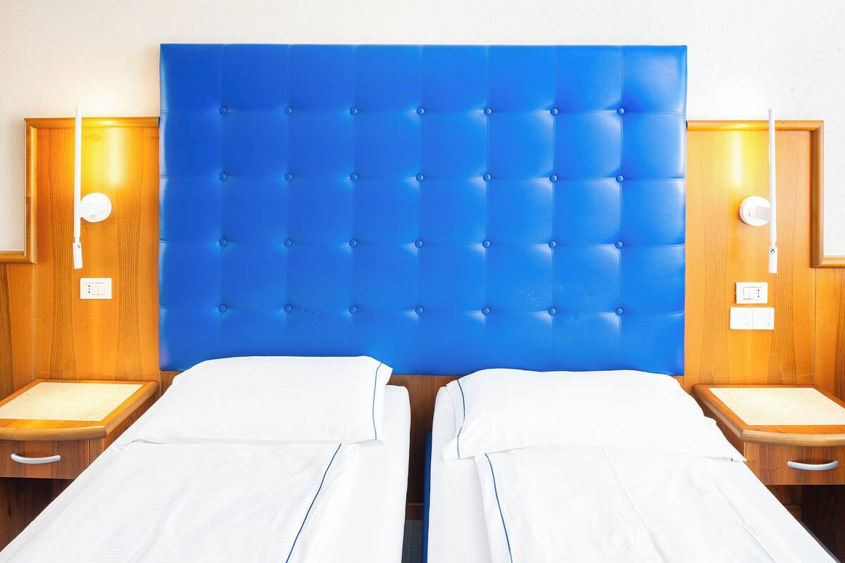 Habitación de hotel con dos camas, cabecera tapizada en azul, mesitas de noche de madera y lámparas fijadas a la pared.