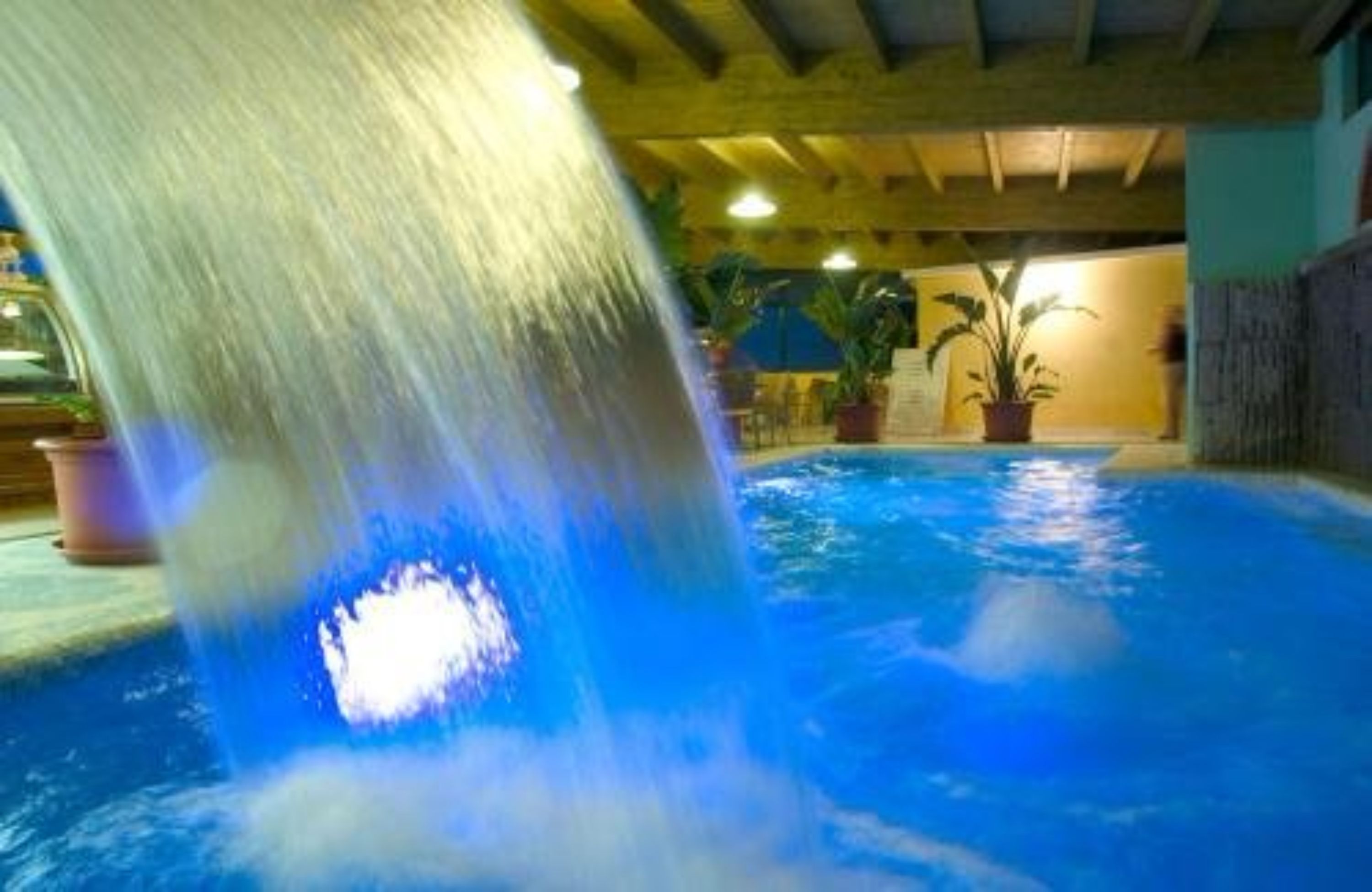 A pool with a waterfall feature, surrounded by potted plants and a wooden ceiling structure.