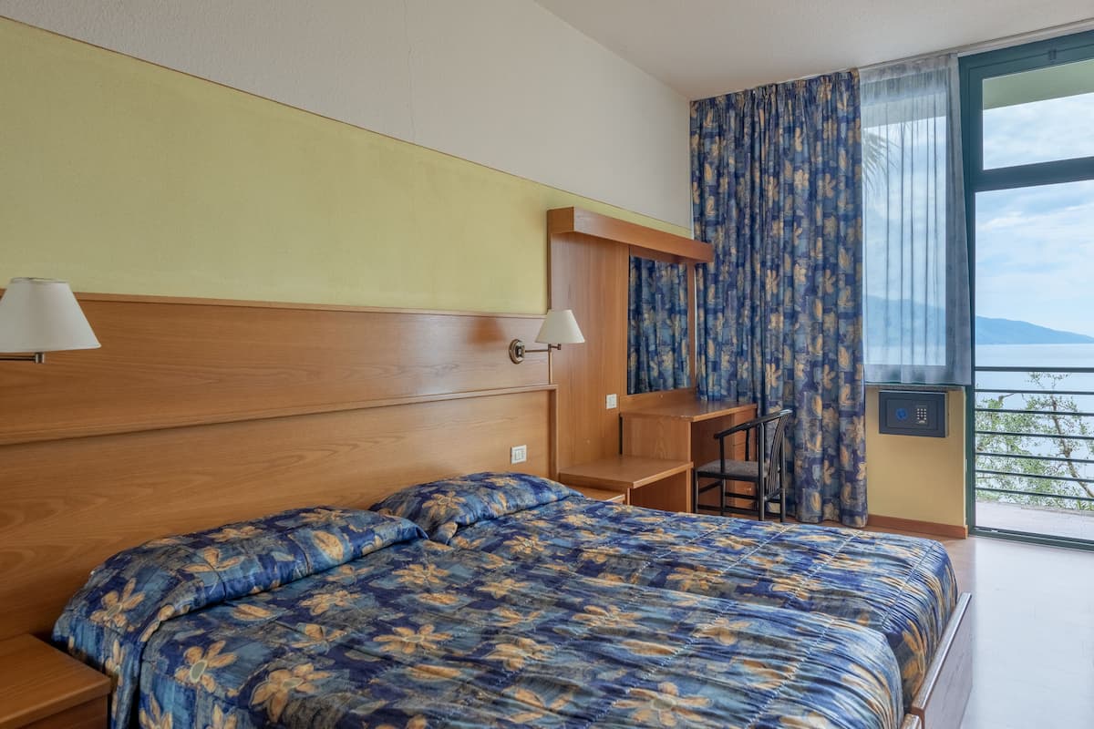 Habitación de hotel con cabecera de madera, cama con colcha estampada, escritorio, dos lámparas y vista al mar a través de un ventanal grande.
