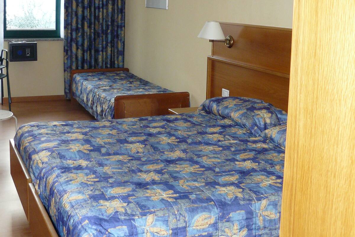 Habitación de hotel con dos camas individuales, una ventana con cortinas, una lámpara de noche y una pequeña mesa con una silla.