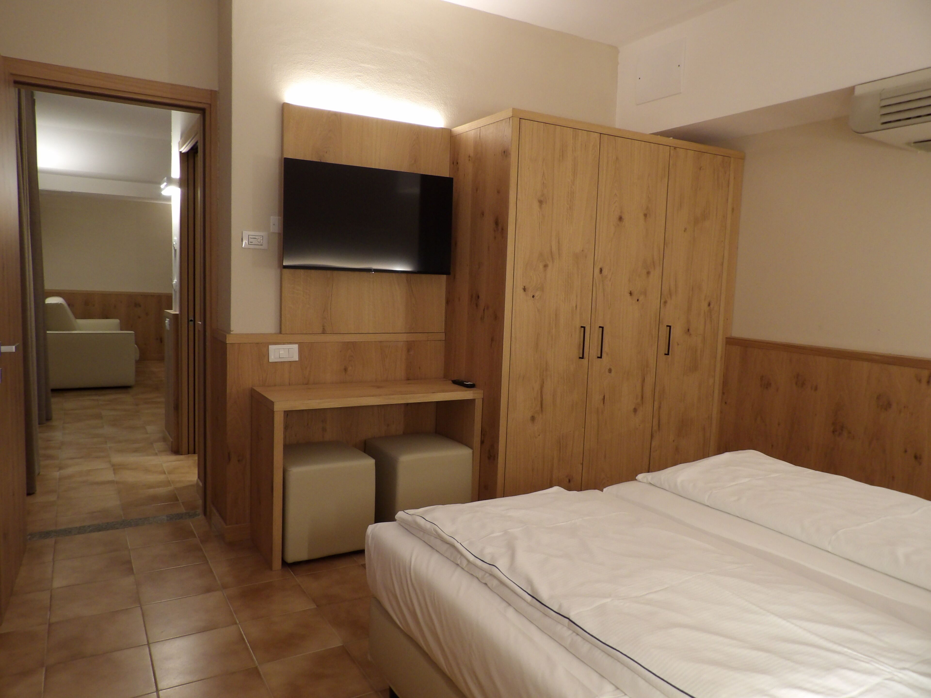 A bedroom with a bed, a TV mounted on the wall, a wooden wardrobe, and a desk with a chair.