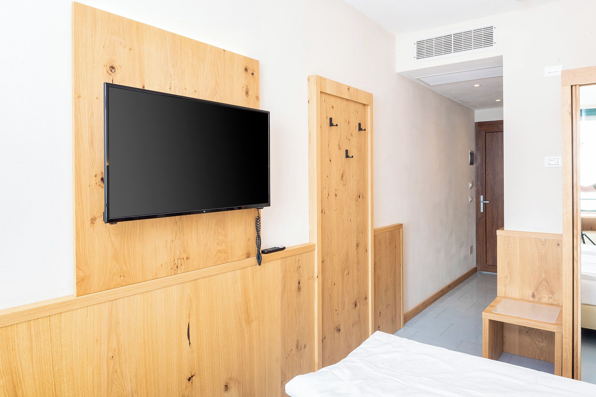 A hotel room with a wooden wall panel, a flat-screen TV mounted on it, a wooden door with a handle, and a small wooden bench.
