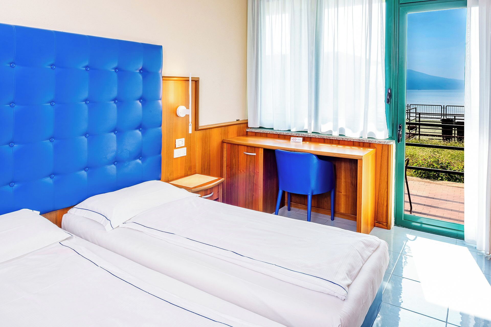 Ein Hotelzimmer mit einem großen Bett, einem Schreibtisch mit Stuhl, einem Fenster mit Meerblick und einem Balkon mit Geländer.