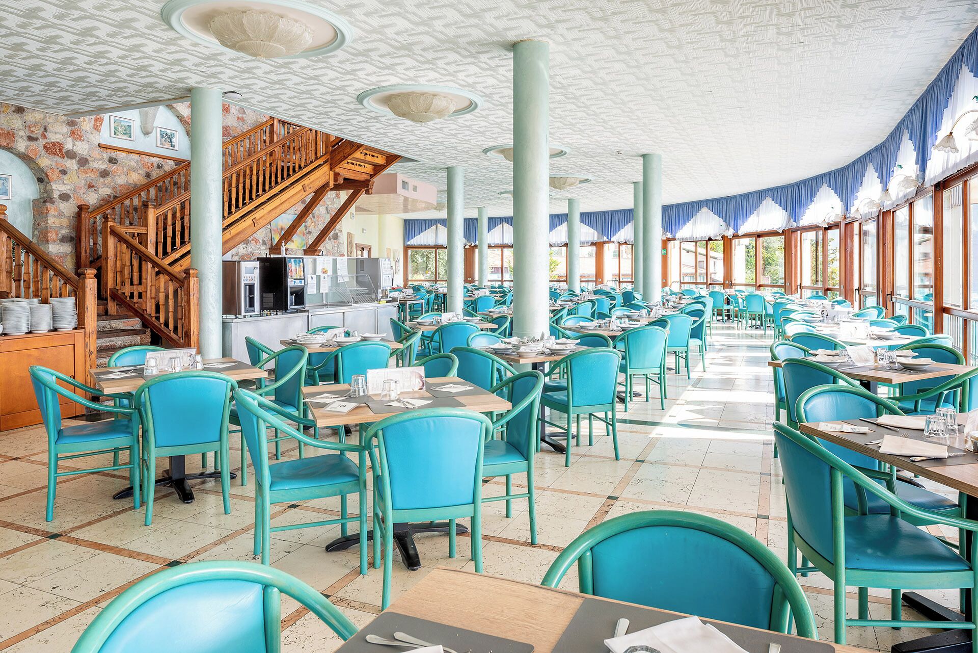 A spacious restaurant with turquoise chairs and tables, a wooden staircase, and a kitchen area.