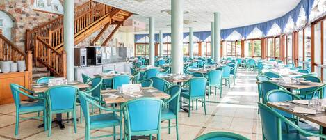 A spacious restaurant with turquoise chairs and tables, a wooden staircase, and a kitchen area.