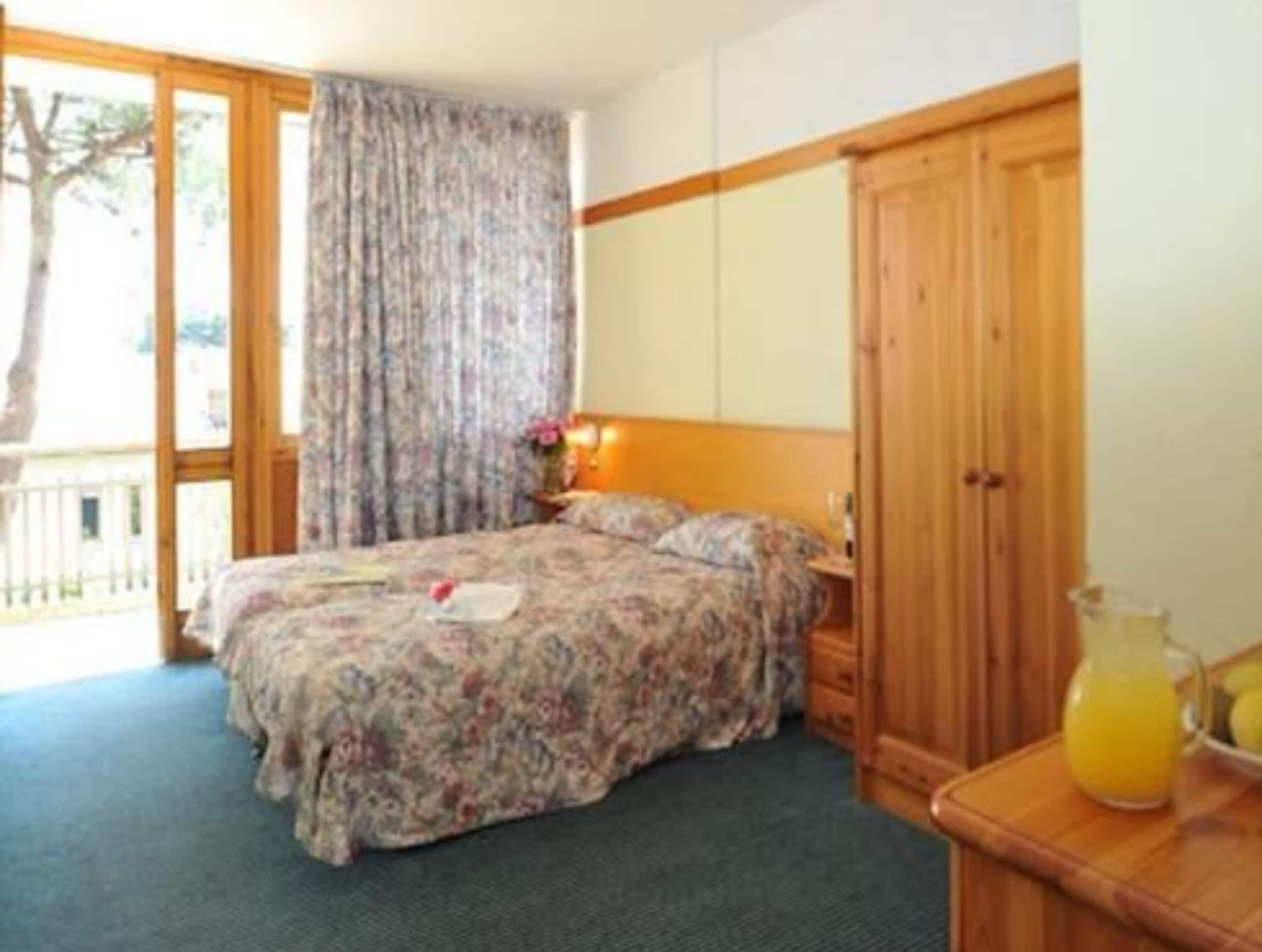 Un dormitorio con cama, mesita de noche, armario de madera, ventana con cortinas y una mesa con jarra de jugo de naranja.