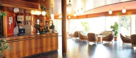 Un hall d’hôtel avec un comptoir d’accueil en bois, plusieurs chaises et une grande fenêtre laissant entrer la lumière naturelle.