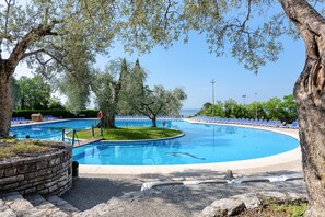 A swimming pool with a stone border, surrounded by trees and blue lounge chairs. - Hotel Marco Polo (Garda)