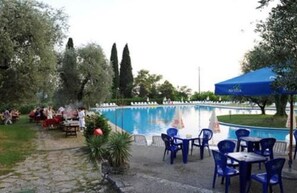 An outdoor pool area with blue chairs and tables, surrounded by trees and a stone pathway. - Hotel Marco Polo (Garda)