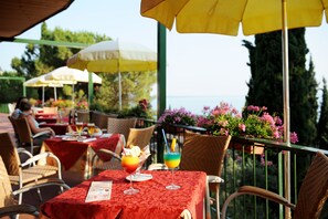 Un área de comedor al aire libre con mesas cubiertas de manteles rojos, sombrillas amarillas y blancas, sillas de mimbre y macetas con flores rosadas.